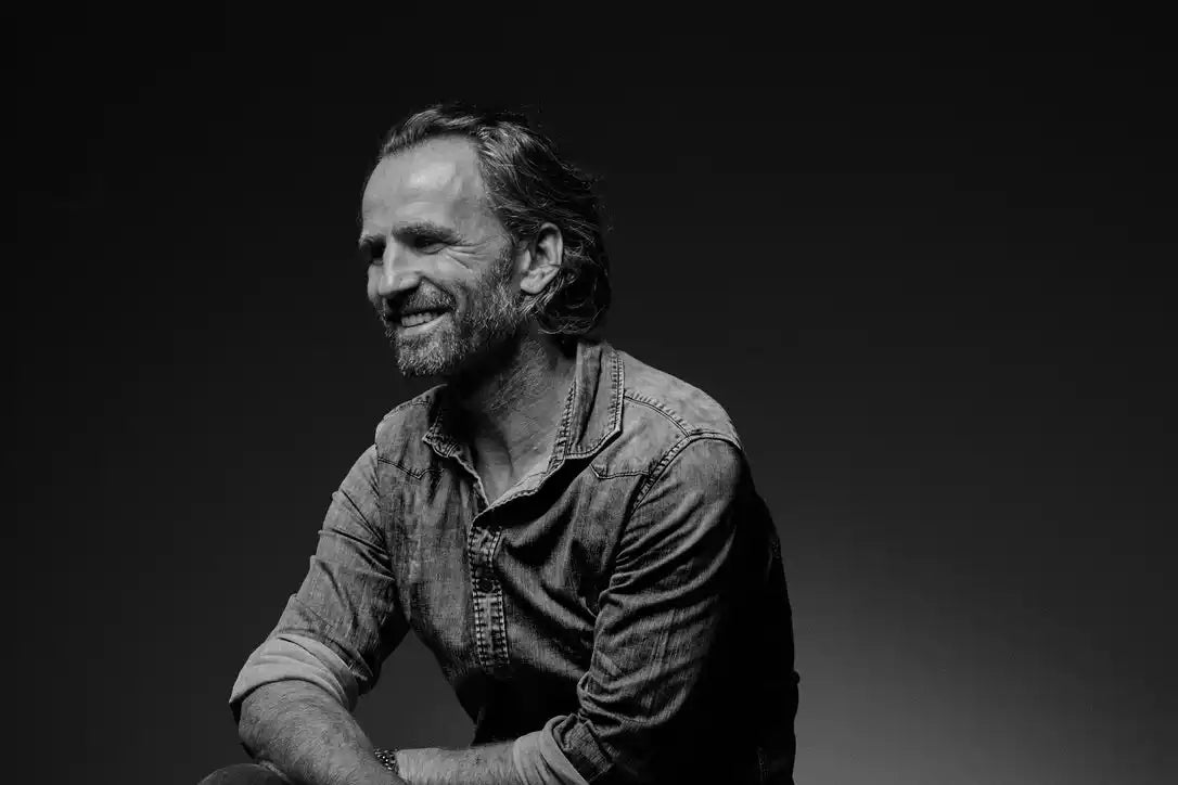 A smiling man with a beard and long hair, wearing a denim shirt.