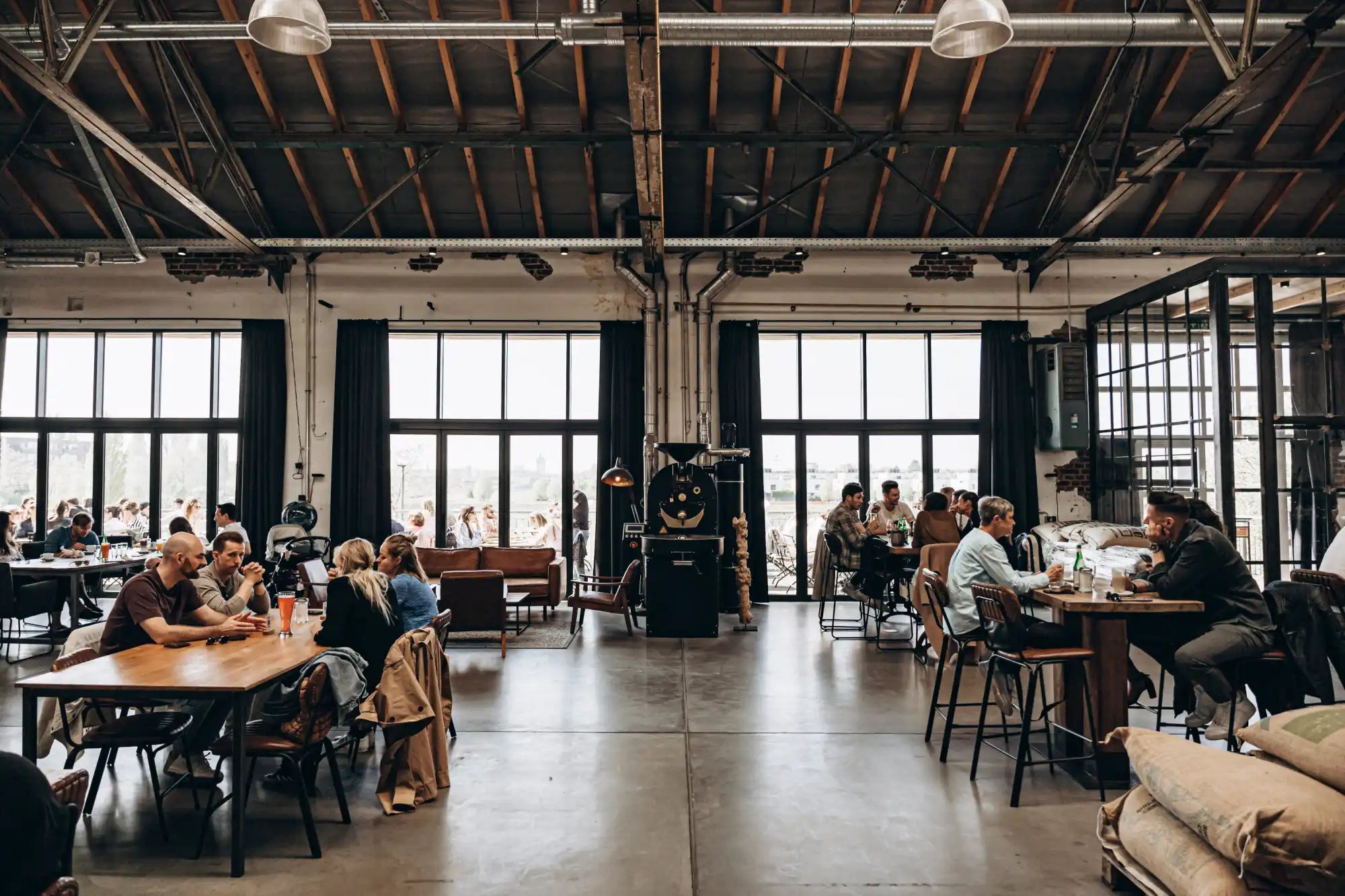 A spacious industrial-style cafe with high ceilings, exposed beams, large windows, and groups of people seated at various tables and lounge areas.